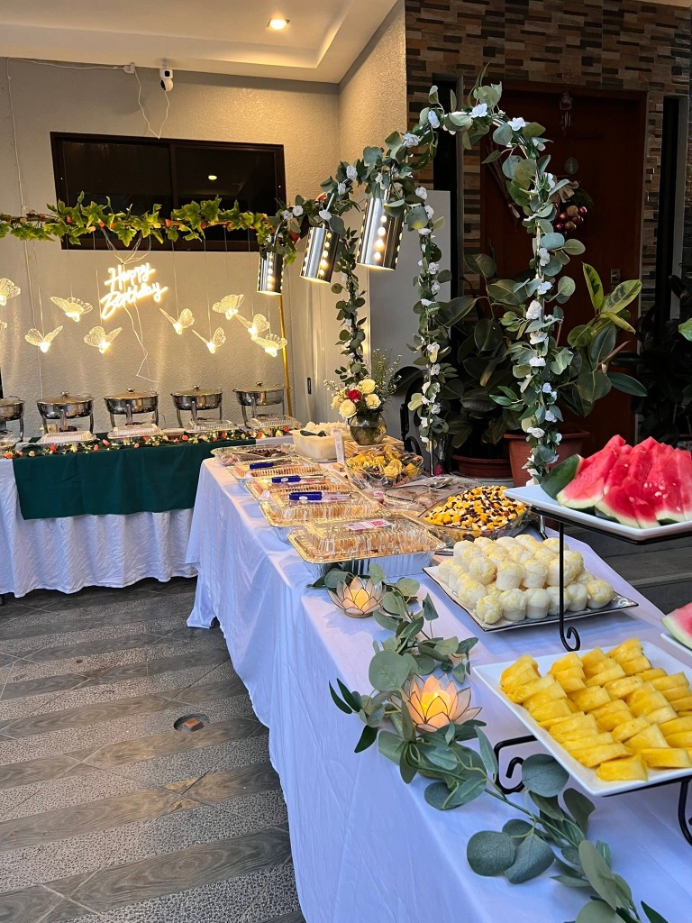 Long dessert table with pastries, puto, fresh fruit platters and greenery beside the main chafing line